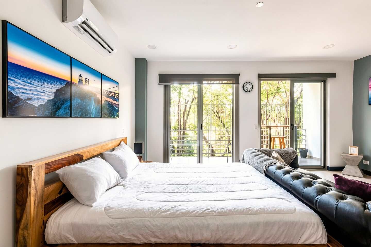 A bedroom with a large bed, a sofa, a view of the outdoors, and a painting on the wall.