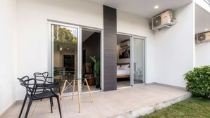 Um pátio moderno ao ar livre com mesa e cadeiras de vidro, uma cama visível pela porta de correr e uma pequena área de jardim.