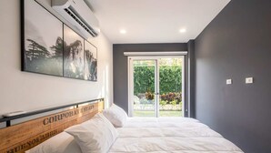 Quarto com cama, ar-condicionado fixado na parede e uma grande janela com vista para área verde.