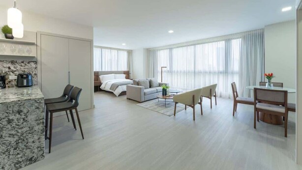 Una habitación de hotel moderna con una isla de cocina de mármol, una cama con cabecera gris, una zona de comedor con mesa y sillas, y grandes ventanales con cortinas translúcidas.