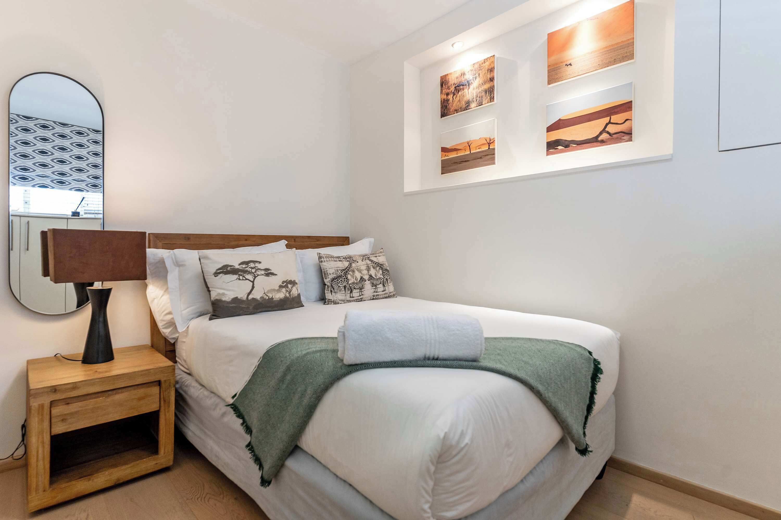 Una cama bien hecha con una manta verde, una lámpara de noche y un espejo sobre una mesita de madera.