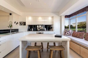 A modern kitchen with a large island, white cabinetry, and a view of a green landscape. - Perfect Views, Sleeps 6, Mouille Point - Emerald Horizon (Cape Town)