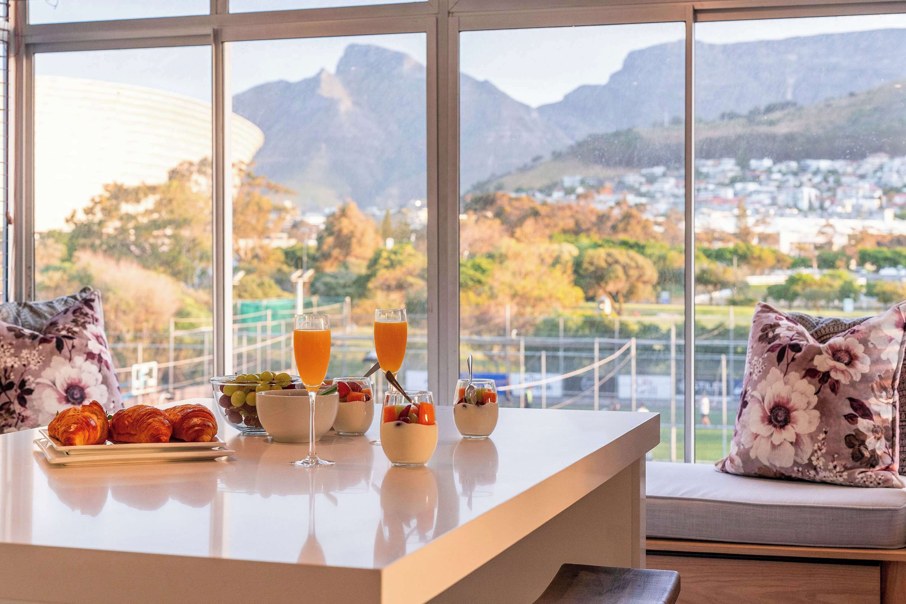 A breakfast table with croissants, fruit, and orange juice, overlooking a scenic mountain view.