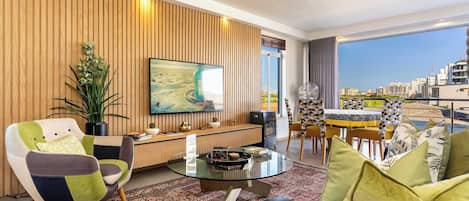 Una sala moderna con un panel de pared de madera, una mesa de centro redonda de vidrio, un sillón colorido, un sofá negro y una zona de comedor con una mesa y sillas geométricas.