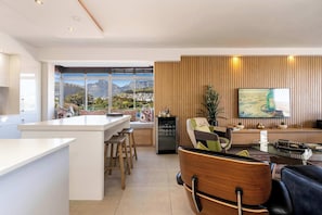 A modern kitchen with a large island, a dining area with a view of mountains, and a living room with a large TV and wooden paneling. - Perfect Views, Sleeps 6, Mouille Point - Emerald Horizon (Cape Town)