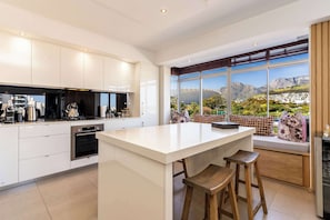 A modern kitchen with a large island, stainless steel appliances, and a view of a mountainous landscape through large windows. - Perfect Views, Sleeps 6, Mouille Point - Emerald Horizon (Cape Town)