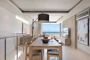 A modern dining area with a long table, chairs, and a view of the sea. - Family Villa, Pool, Ocean Views - Camps Bay Grace (Cape Town)