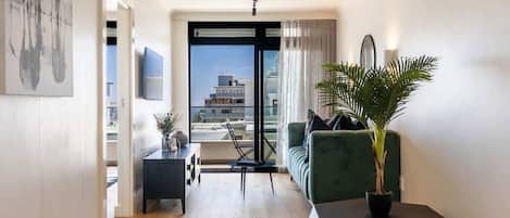 A modern living room with a sofa, a coffee table, and a potted plant. There is a view of buildings through a glass door.