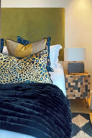 Ein Bett mit gemusterten Kissen, einer schwarzen Tagesdecke, einem Holfnachtkästchen mit einer Lampe und einem geometrischen Teppich.
