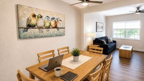 Uma área de refeições com uma mesa de madeira, cadeiras, um laptop, uma xícara de café e uma planta. Há um quadro com pássaros na parede, um ventilador de teto e um sofá ao fundo.