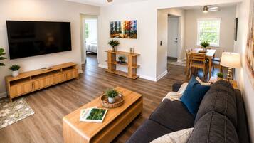 Ein modernes Wohnzimmer mit Flachbildfernseher, Holzmöbeln, Deckenventilator und einem Essbereich mit Tageslicht.