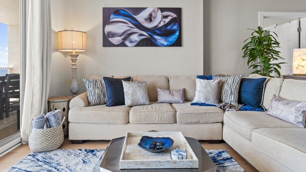 Ein Wohnzimmer mit einem beigen Sofa, einem Couchtisch mit Fernbedienungen, einer Lampe und einem Gemälde an der Wand.