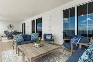 A spacious balcony with a wooden table, blue upholstered sofas, and a large window offering a view of the sky. - Fall Deals + Snowbird Rates | Gulf Front Beach Escape (Panama City Beach)