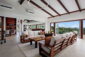 A spacious living area with a large sofa set, a wooden coffee table, and a dining table. There is a view of the outdoors through large windows. - Onomea Oasis (Papaikou)