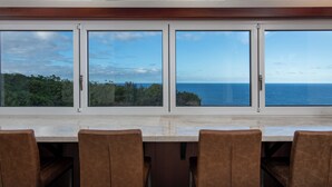 Ein Essbereich mit großem Tisch und Stühlen, große Fenster mit Blick auf das Meer und viel Grün.