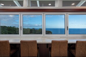 A dining area with a large table and chairs, large windows with a view of the ocean and greenery. - Onomea Oasis (Papaikou)