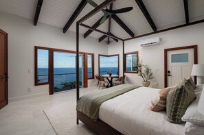 A bedroom with a large bed, a dining table, and chairs, a ceiling fan, and a view of the ocean. - Onomea Oasis (Papaikou)