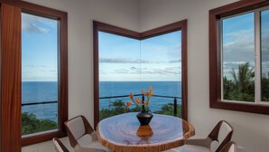 Ein runder Holztisch mit einer Vase orangener Blumen, umgeben von vier Stühlen, in einem Raum mit großen Fenstern und Meerblick.