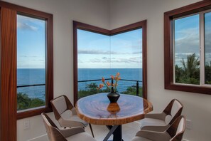 A round wooden table with a vase of orange flowers, surrounded by four chairs, in a room with large windows offering a view of the sea. - Onomea Oasis (Papaikou)