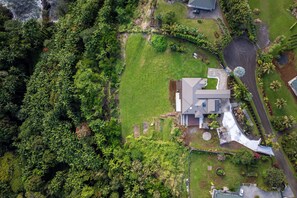 An aerial view of a modern house surrounded by lush greenery, with a well-maintained lawn and a driveway leading to the entrance. - Onomea Oasis (Papaikou)