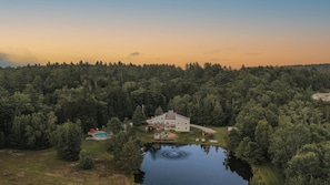 A large house with a deck and a swimming pool, surrounded by dense forest and a lake.