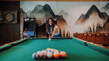 A pool table with a person playing, surrounded by arcade machines and a mural of mountains.