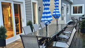 Una terraza con una mesa y sillas de metal, dos sombrillas azuladas a rayas y una casa de fachada gris.