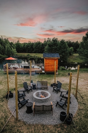 Un espace circulaire aménagé avec un foyer central, entouré de chaises et de tables, une terrasse en bois avec des fauteuils de jardin, et une petite cabane en arrière-plan.