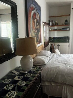 A hotel room with a bed, a lamp, a mirror, a painting, and a shelf with books and decorative items. - Los Cabos Retreat
3/3 home, 24 Hr. security, private pool gated compound (San José del Cabo)
