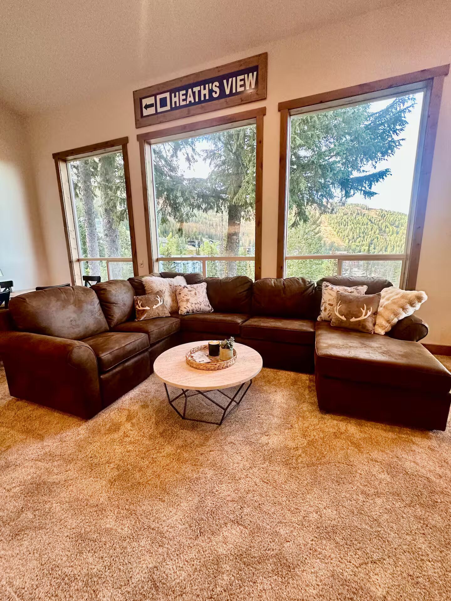 A living room with a sectional sofa, a round coffee table, and large windows offering a view of a forested landscape.