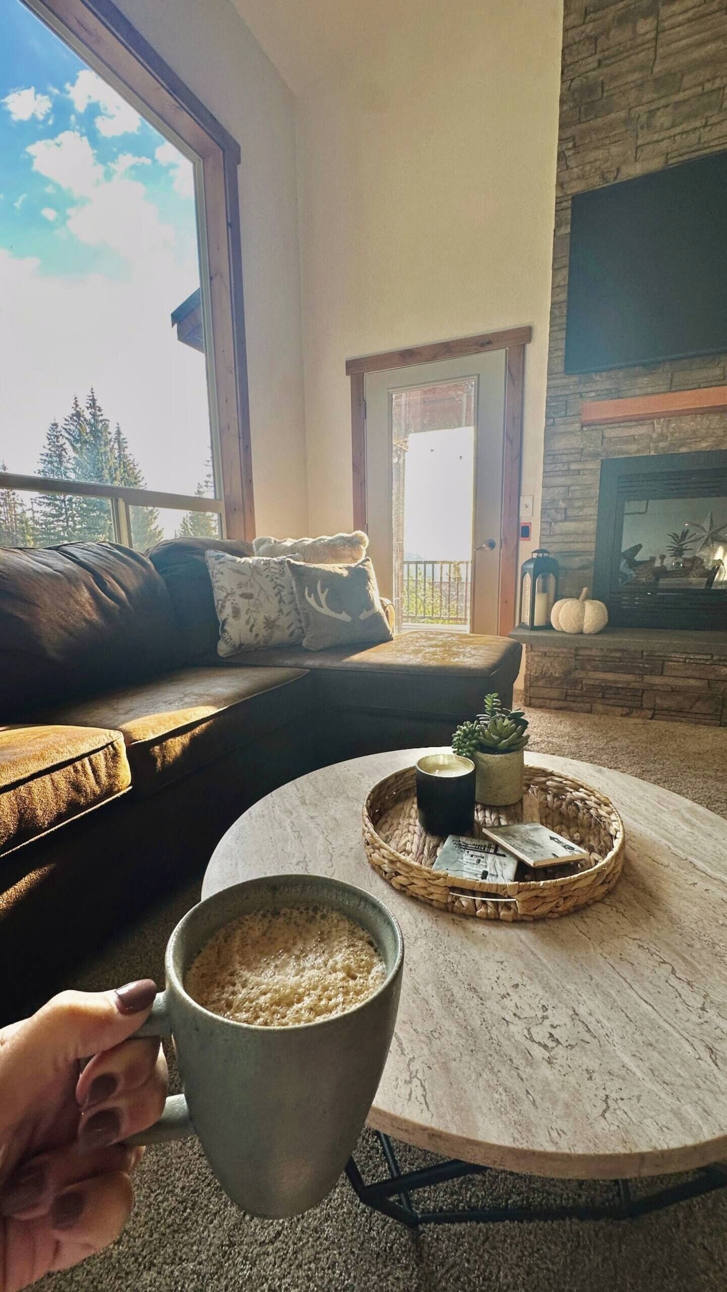 A cozy living room with a leather sofa, a round coffee table with a mug, a fireplace, and a view of the outdoors.