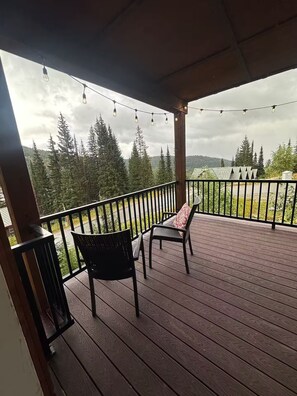 Um deck coberto ao ar livre com duas cadeiras e uma mesa, com vista para uma paisagem arborizada.