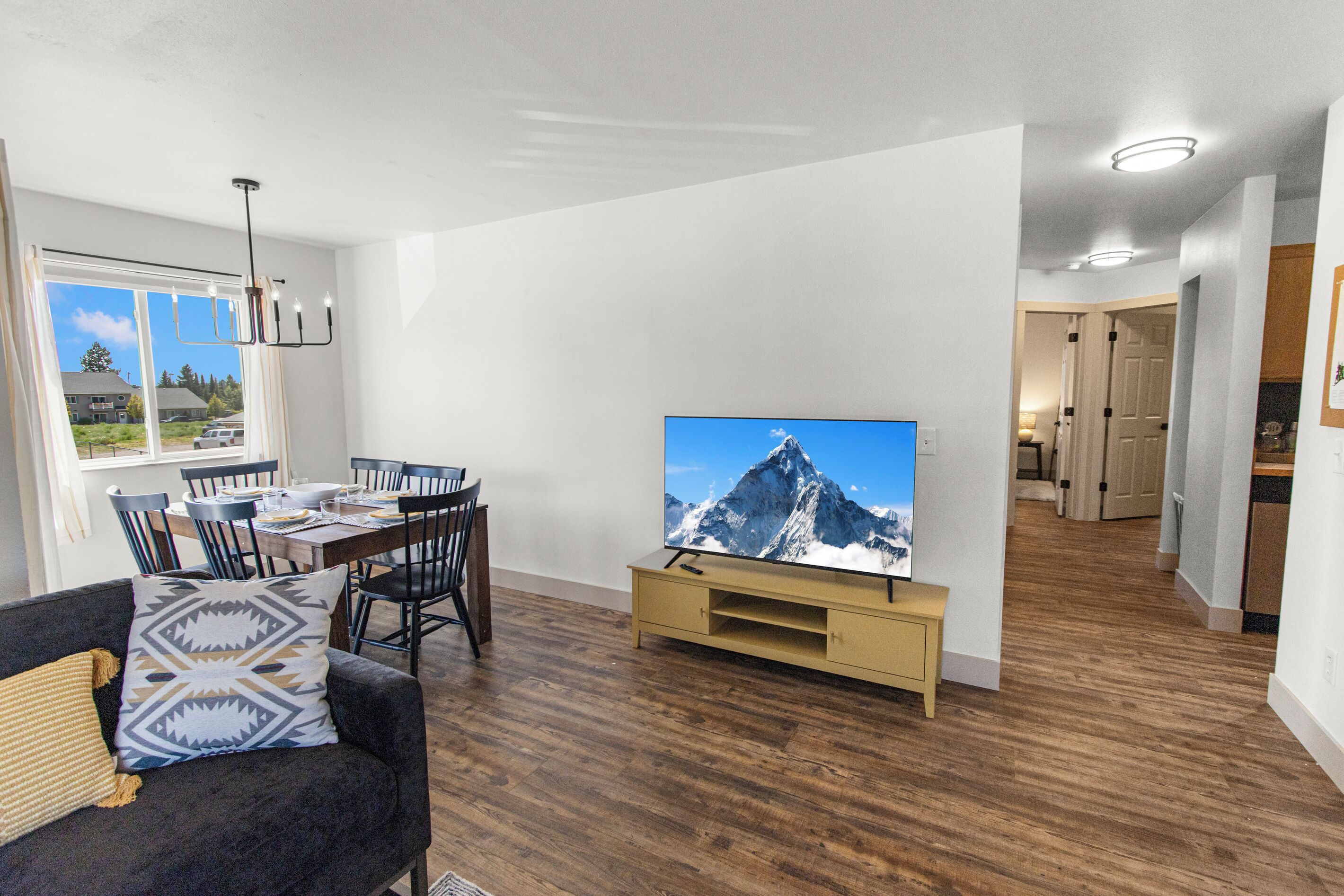 A modern living room with a flat-screen TV, dining area, and a hallway leading to a kitchen.