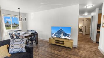 A modern living room with a flat-screen TV, dining area, and a hallway leading to a kitchen.