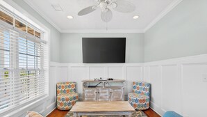 A room with a ceiling fan, a flat-screen TV mounted on the wall, a wooden coffee table, floral-patterned armchairs, and a sofa with a patterned rug. - Spacious 7-bedroom Beach House in fantastic Ocean Isle Beach w/ Private Pool&AC (Ocean Isle Beach)