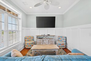 Ein Zimmer mit Deckenventilator, einem an der Wand befestigten Flachbildfernseher, einem Holztisch, floralen Ohrensesseln und einem Sofa mit gemustertem Teppich.
