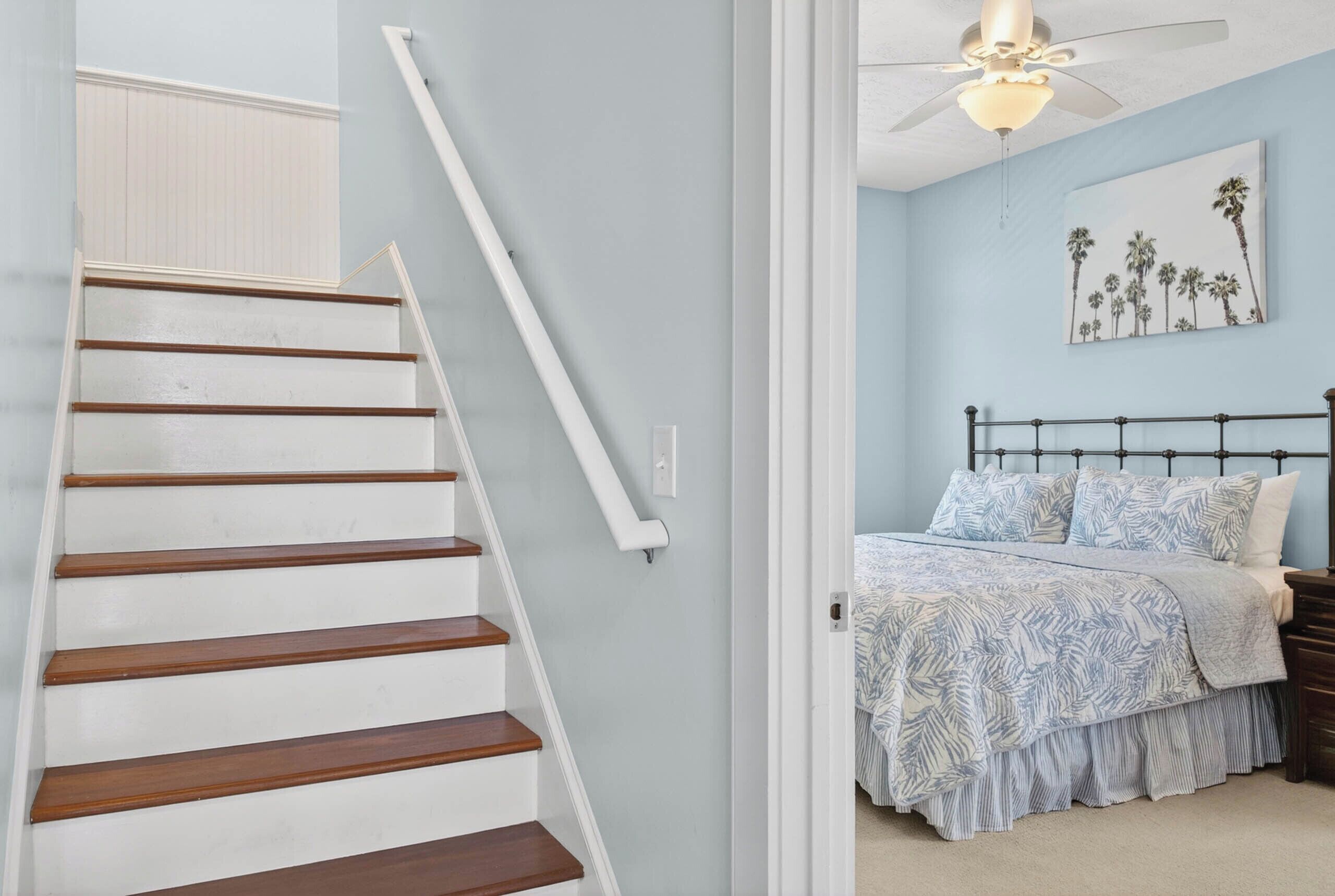 A staircase with wooden steps and white handrails, leading to a bedroom with a bed, a nightstand, and a ceiling fan.