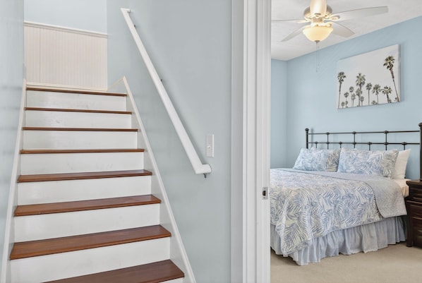 Una escalera con escalones de madera y barandillas blancas, que conduce a un dormitorio con cama, mesita de noche y ventilador de techo.
