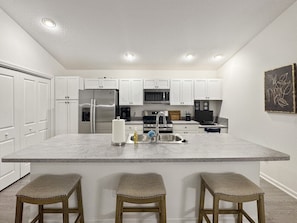 A kitchen with white cabinets, a large island with stools, stainless steel appliances, and recessed ceiling lights.