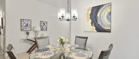 Comedor con mesa de vidrio redonda, cuatro sillas, una araña y cuadros en la pared.