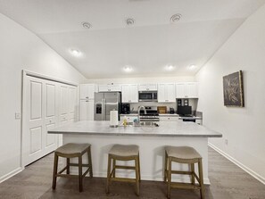 Una cocina con armarios blancos, un refrigerador de acero inoxidable, un microondas y un fregadero. Hay tres taburetes de madera en la isla.