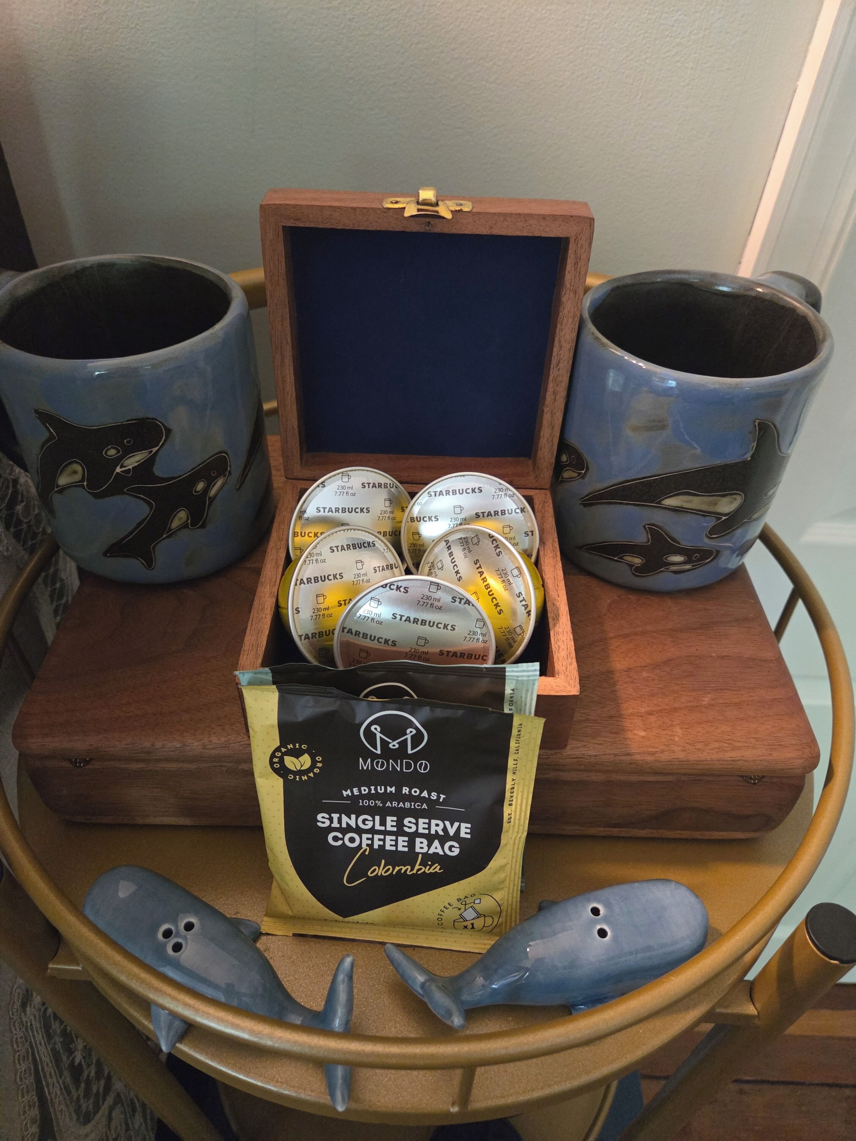Una caja de madera que contiene un paquete de café, cápsulas de café y dos tazas de café con diseños de ballena.