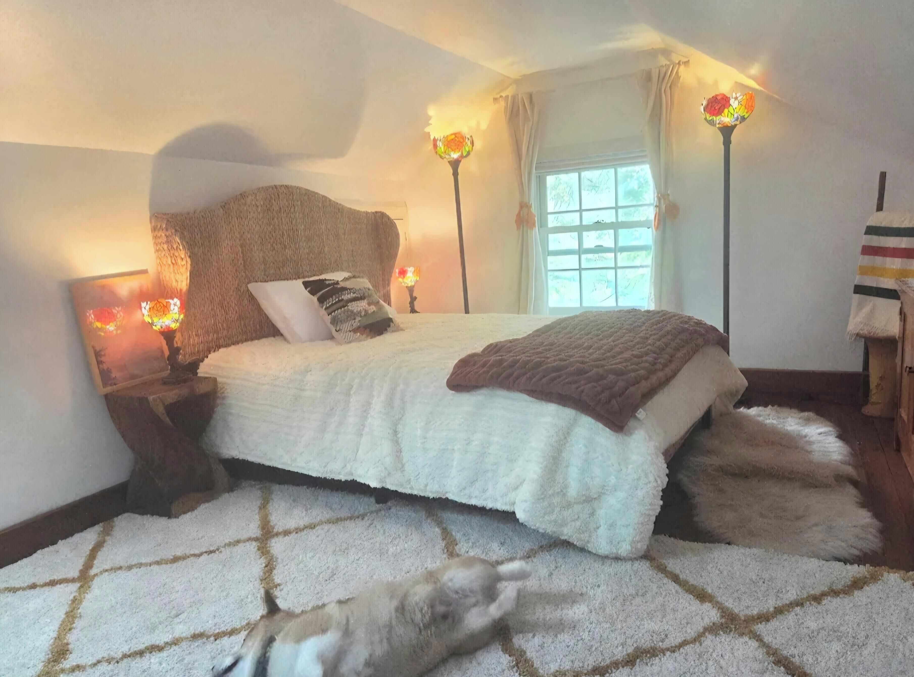 A bedroom with a large bed, two bedside lamps, a window with curtains, and a fluffy rug.
