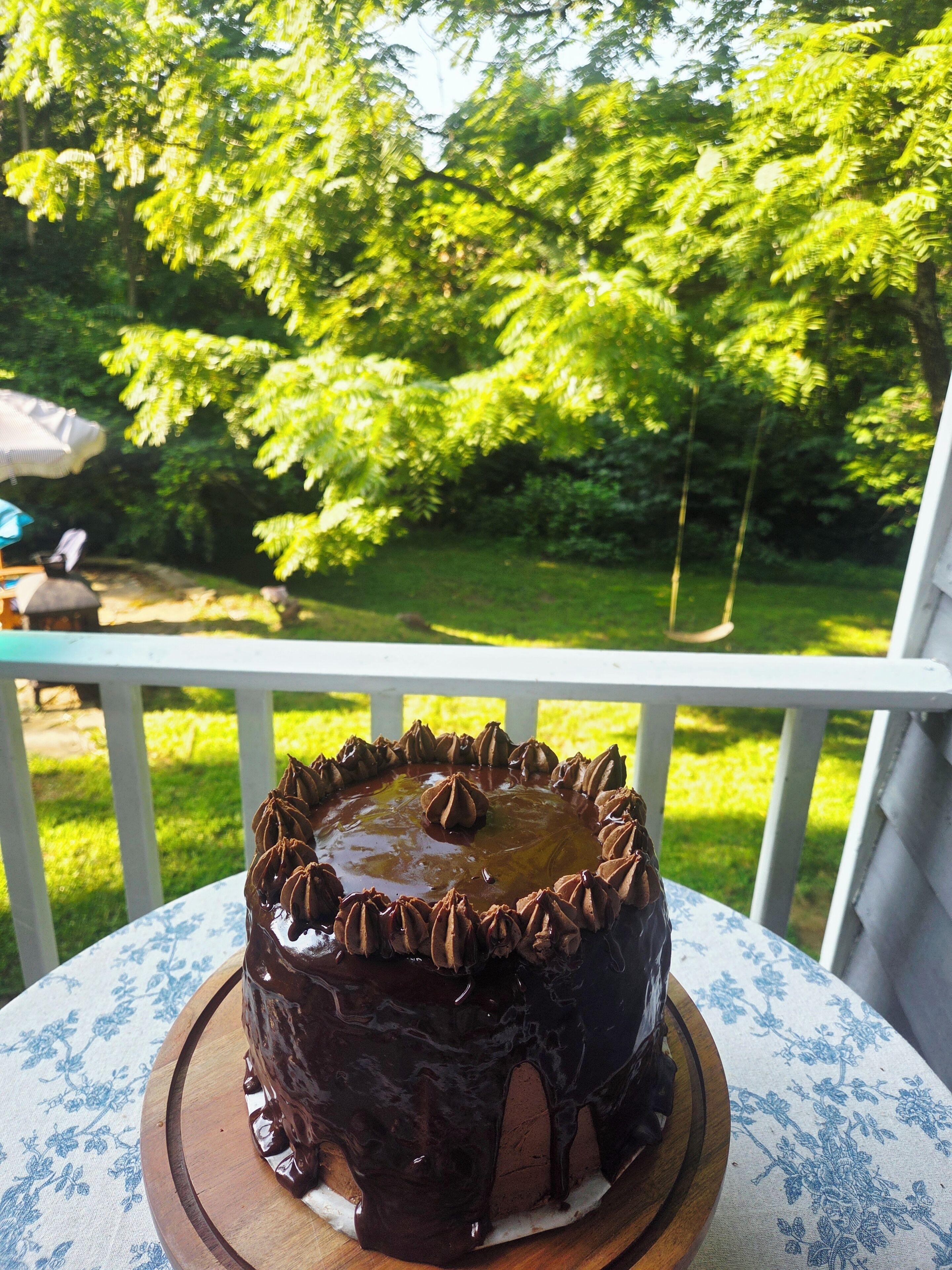 Um bolo de chocolate com cobertura brilhante sobre uma tábua de madeira, colocado em uma mesa com toalha de mesa florida.