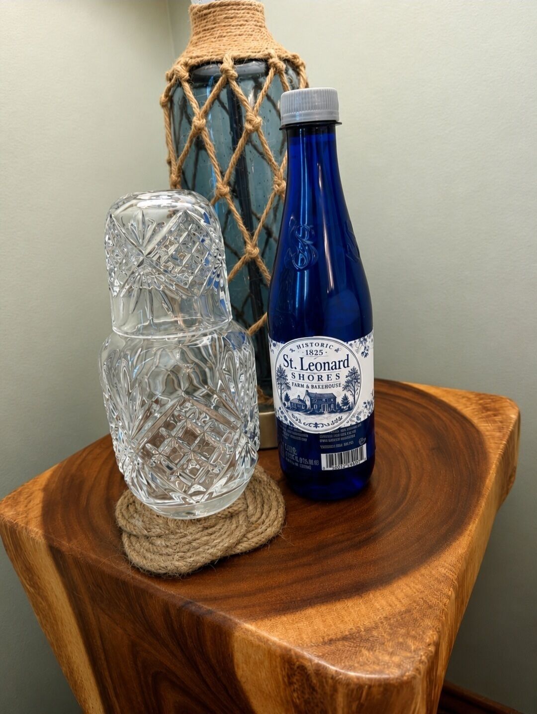 Una botella de vidrio transparente con etiqueta azul, un florero de vidrio transparente con diseño de cuerda y una superficie de madera.