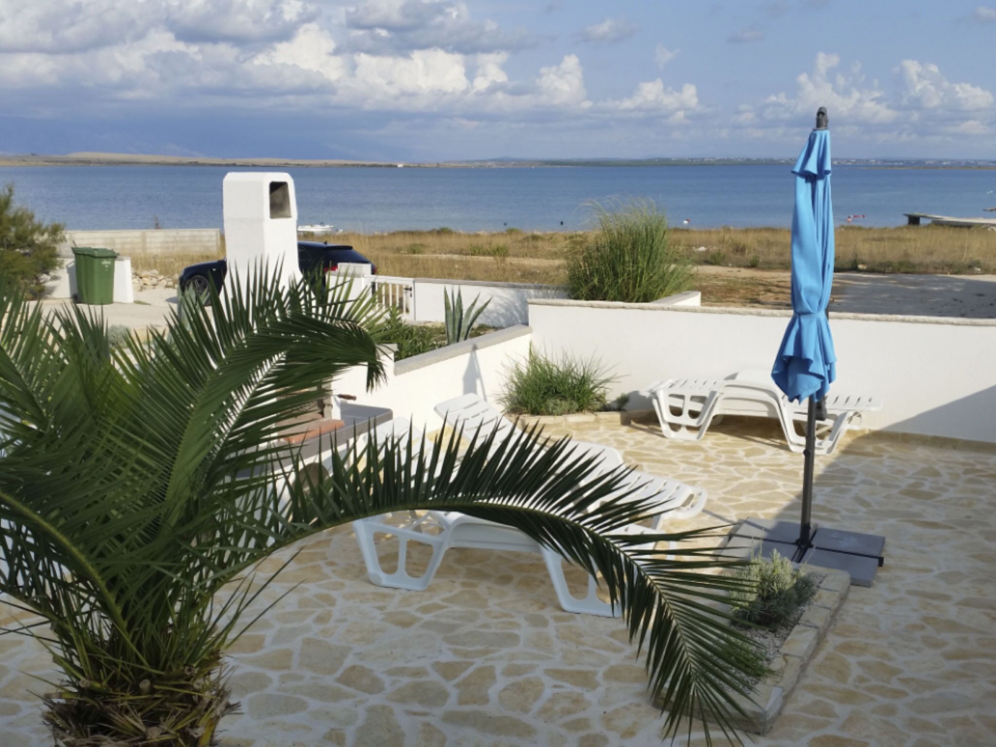 A patio with stone flooring, white furniture, a blue umbrella, and a view of the sea.