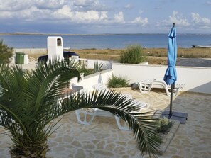 Eine Terrasse mit Steinfliesen, weißen Möbeln, einem blauen Sonnenschirm und Blick aufs Meer.