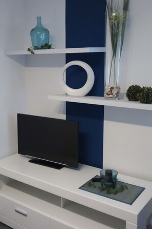Televisão de tela plana em um móvel branco com uma parede azul de destaque, um objeto circular branco e um vaso com plantas.