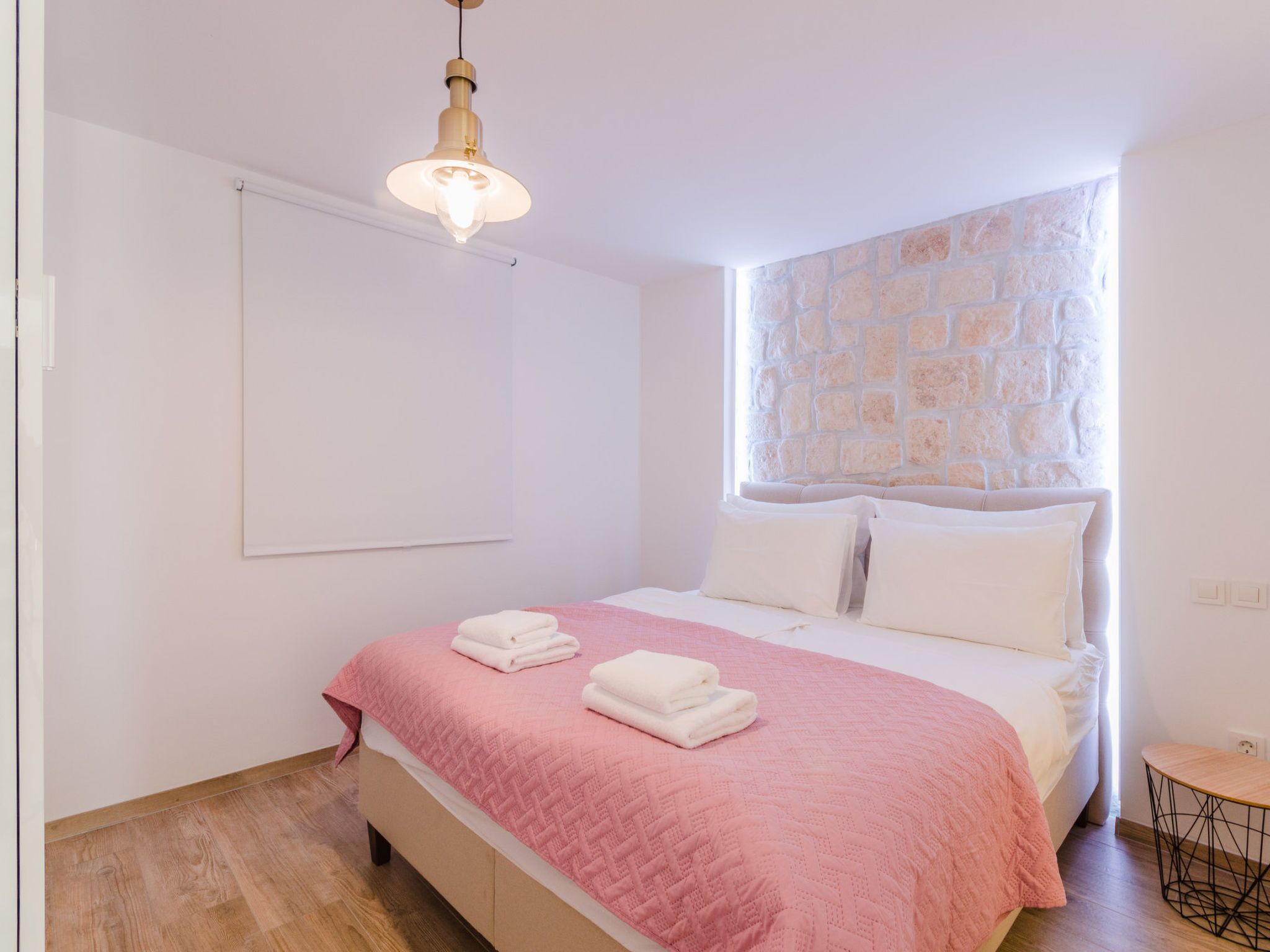 A bedroom with a bed, a window with a stone wall, a hanging light fixture, and a small round table.