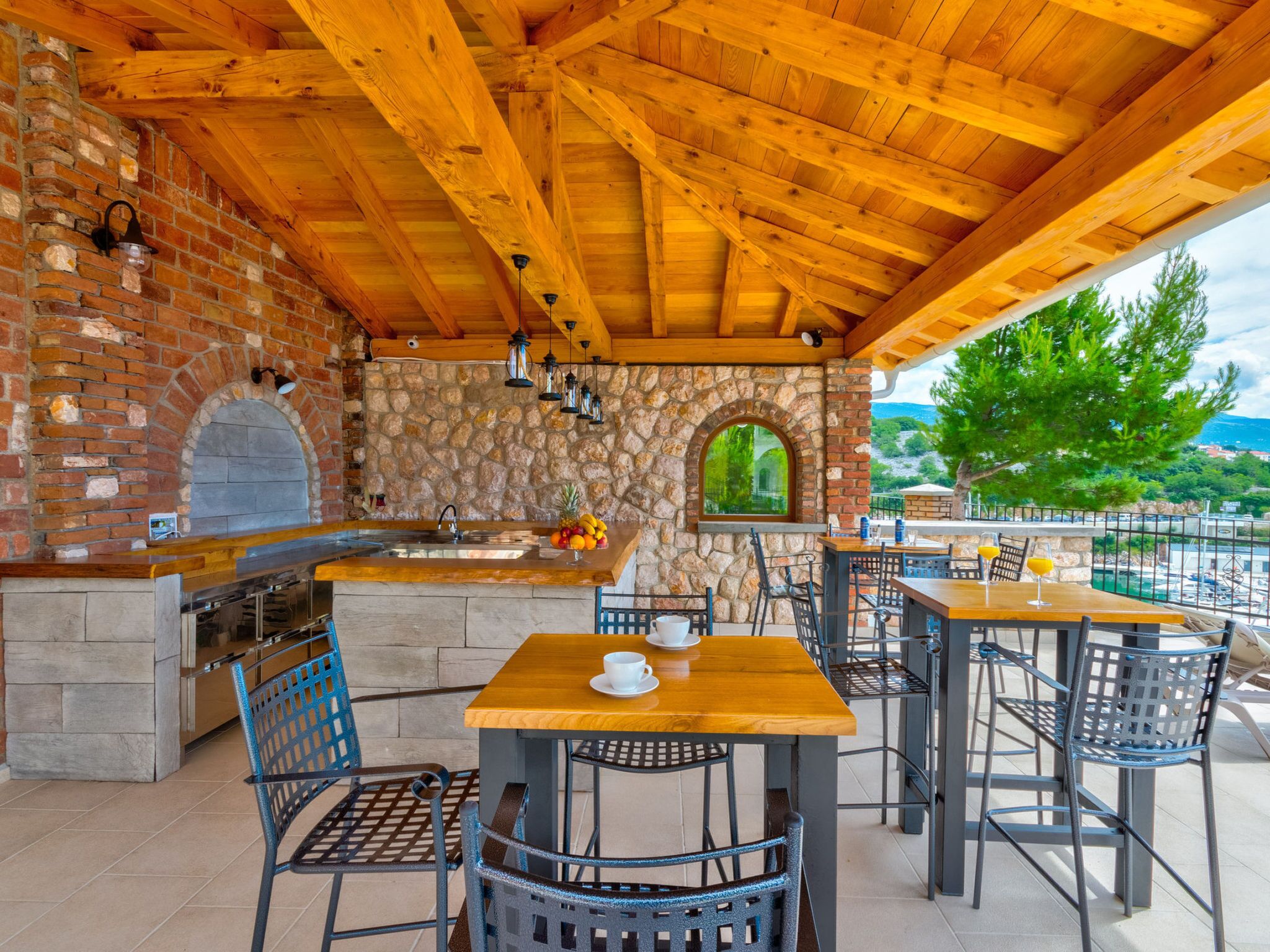 An outdoor patio with a brick oven, stone wall, wooden ceiling, and outdoor seating.